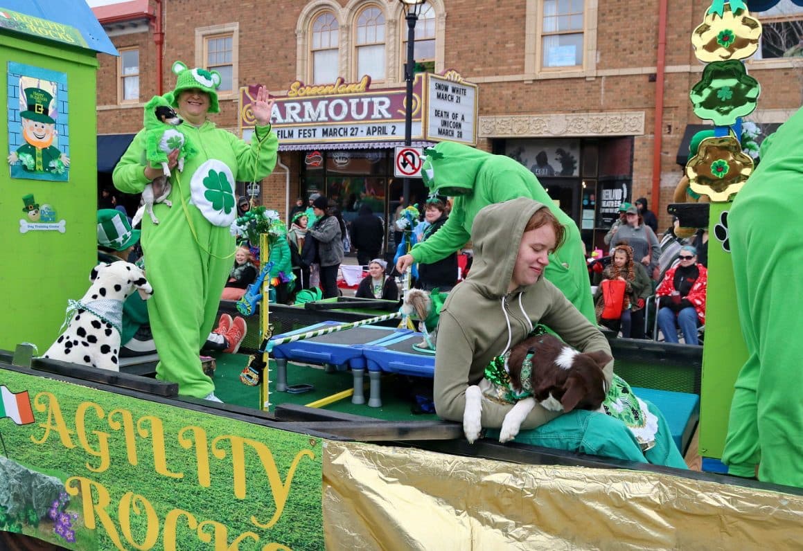 A charity float in green with costumed riders rolls past the Armour Theatre