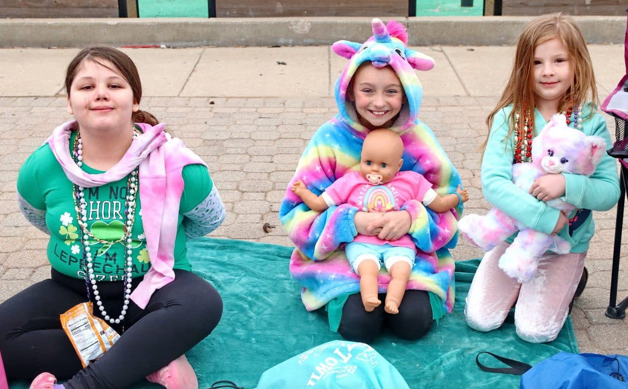 Three girls in rainbow and green wait at the curb for the parade