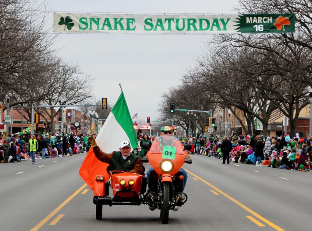 A rider with an Irish flag on an orange sidecar motorcycle leads the parade under the Snake Saturday banner
