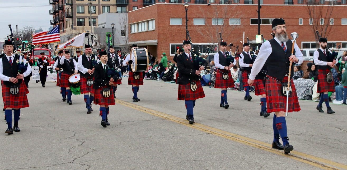 A pipe and drum band in kilts marching down the parade route past the crowd